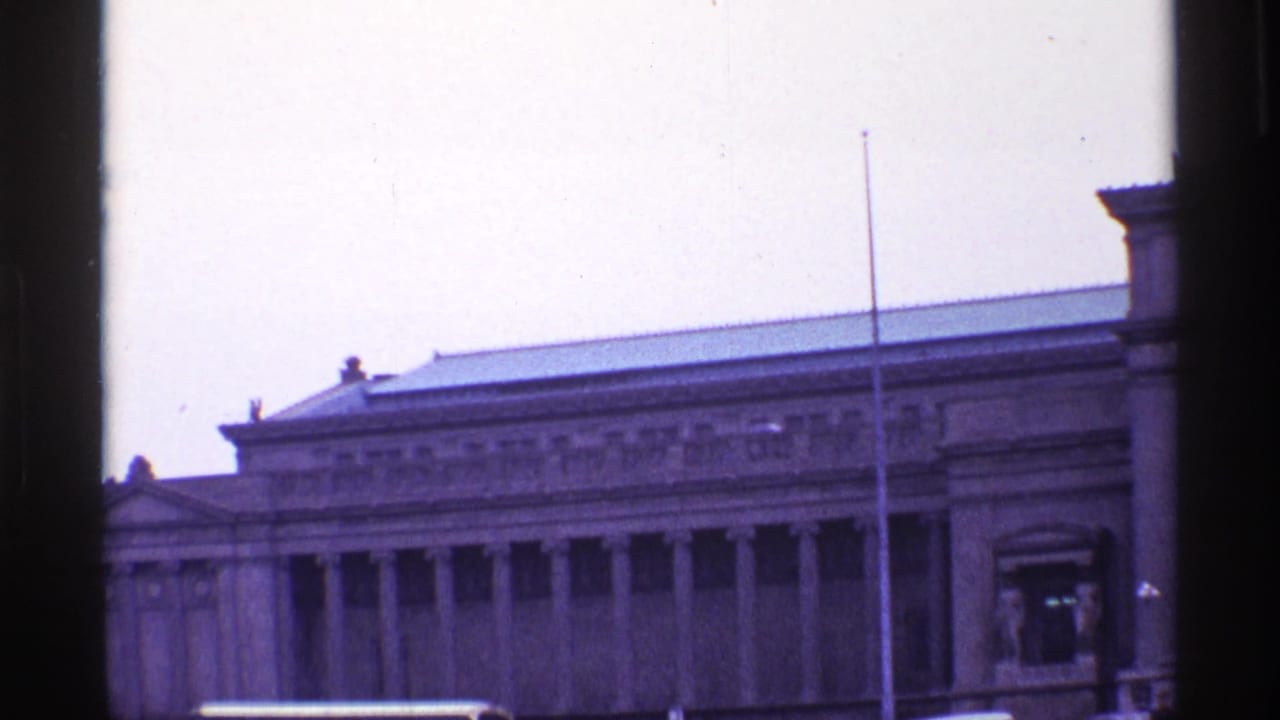 1968 Chicago home-movie frame showing a grand columned building with flagpole, street traffic, and dark foreground window edges.