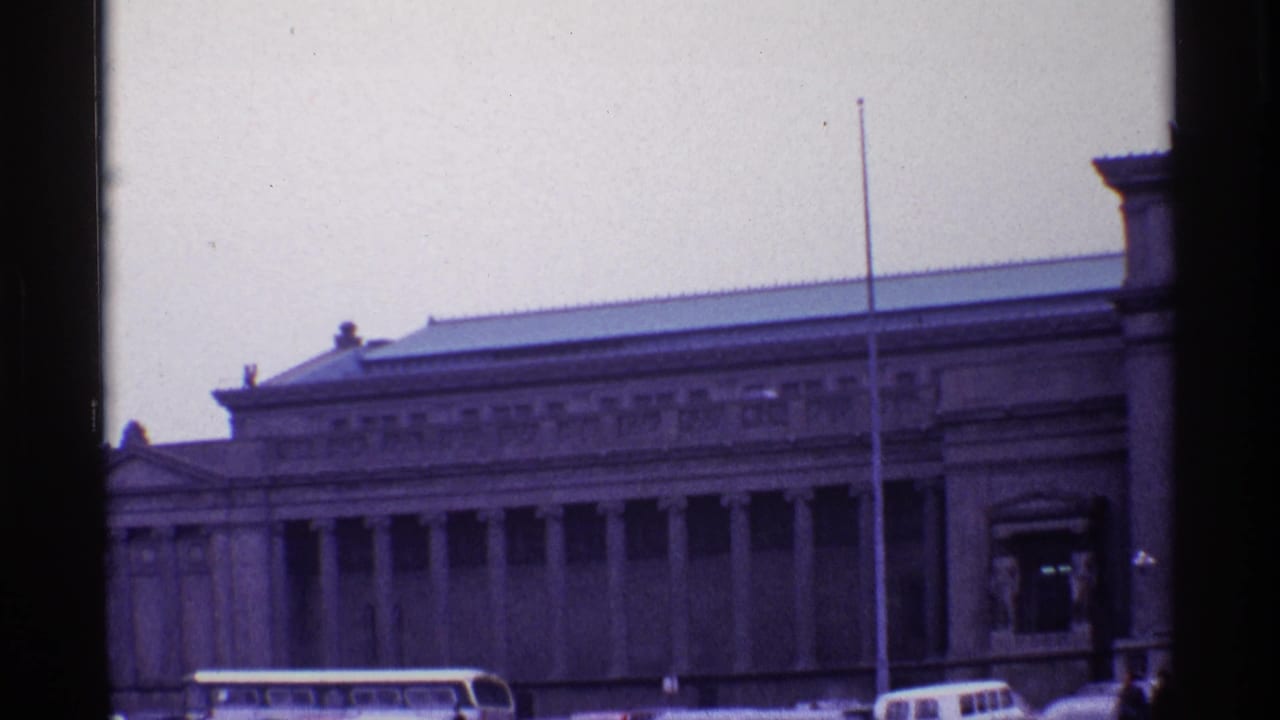 Similar view of the columned building with a flagpole, bus, and cars partly visible below.