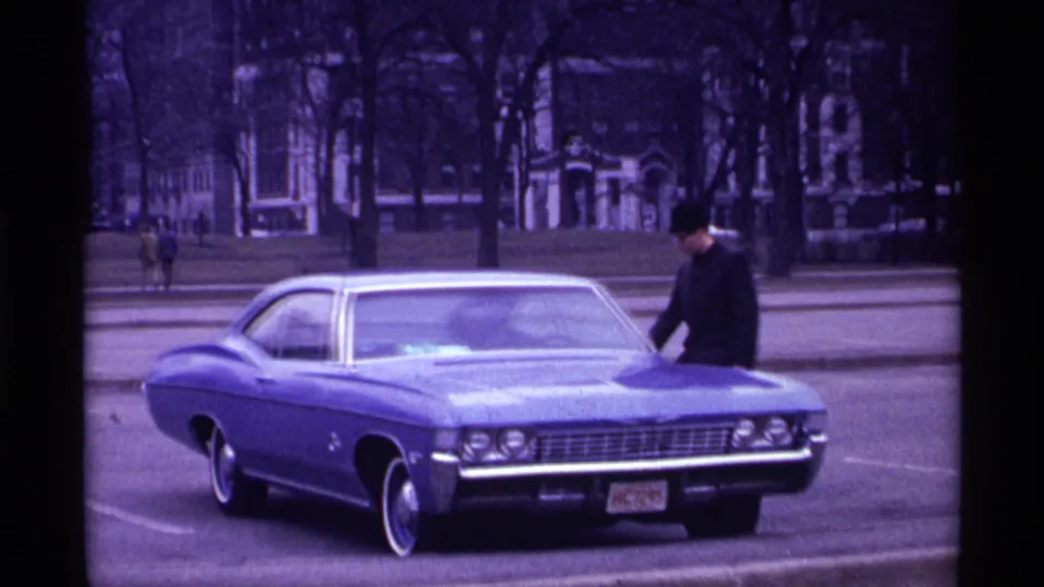 1968 Chicago home-movie frame with a man beside a blue late-1960s car, trees and buildings in the background.