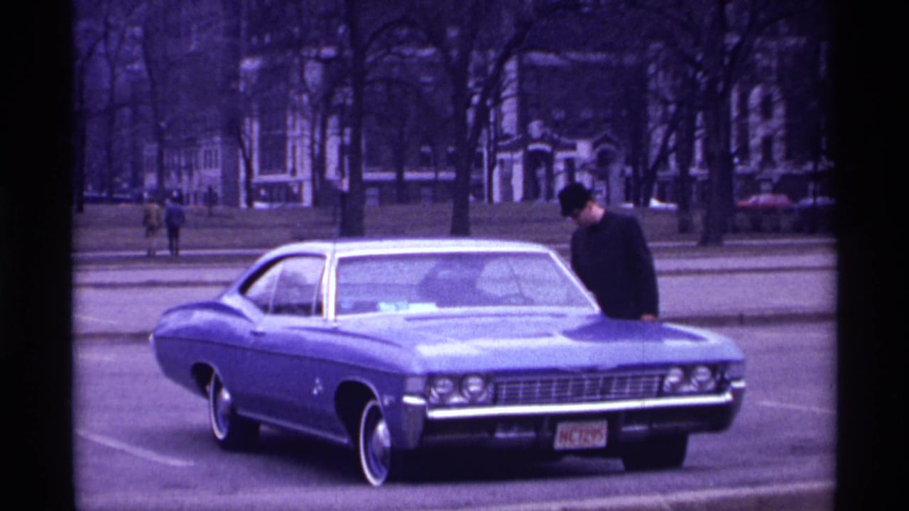 Blue late-1960s car parked on a city street as a man leans near the hood.