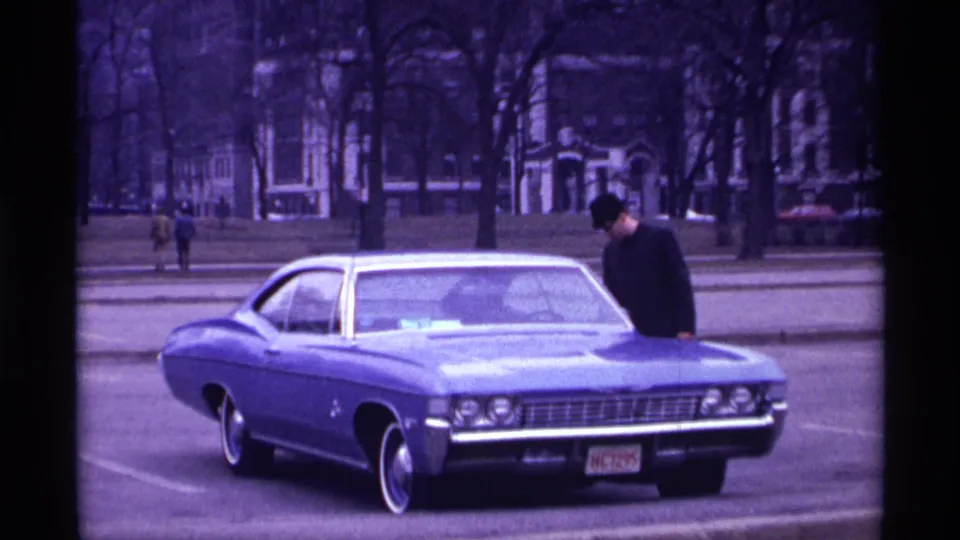 In 1968 Chicago home-movie footage, a man in a dark jacket stands by the hood of a blue Chevrolet Impala on a city street.