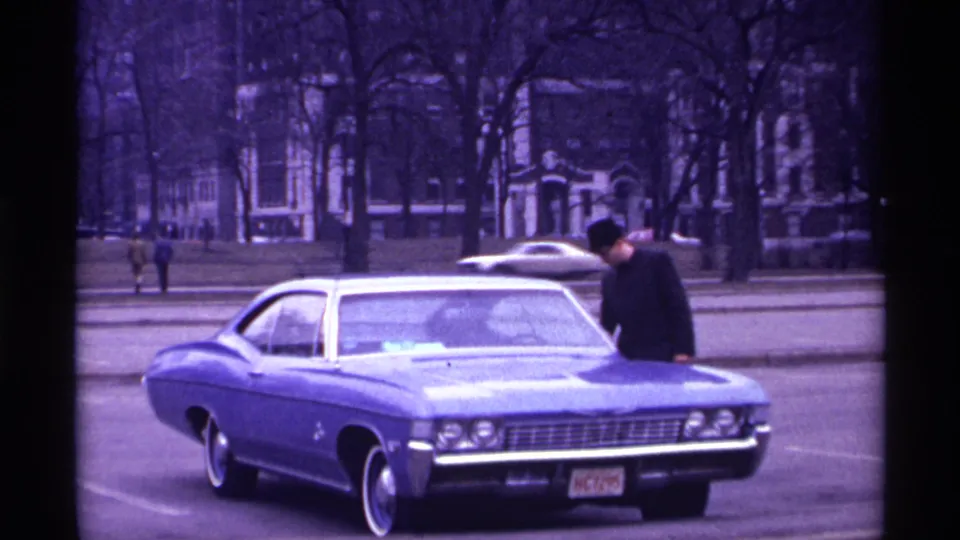 The man remains near the hood of the blue Chevrolet Impala while a light-colored car passes in the Chicago street background.