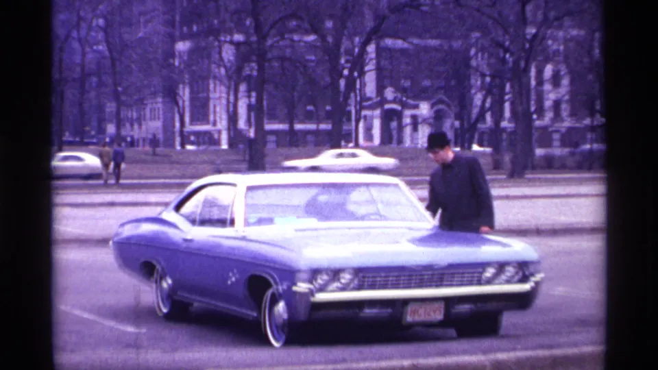 A blue 1968 Chevrolet Impala sits parked in Chicago as the man leans over the hood, with distant pedestrians and traffic visible.