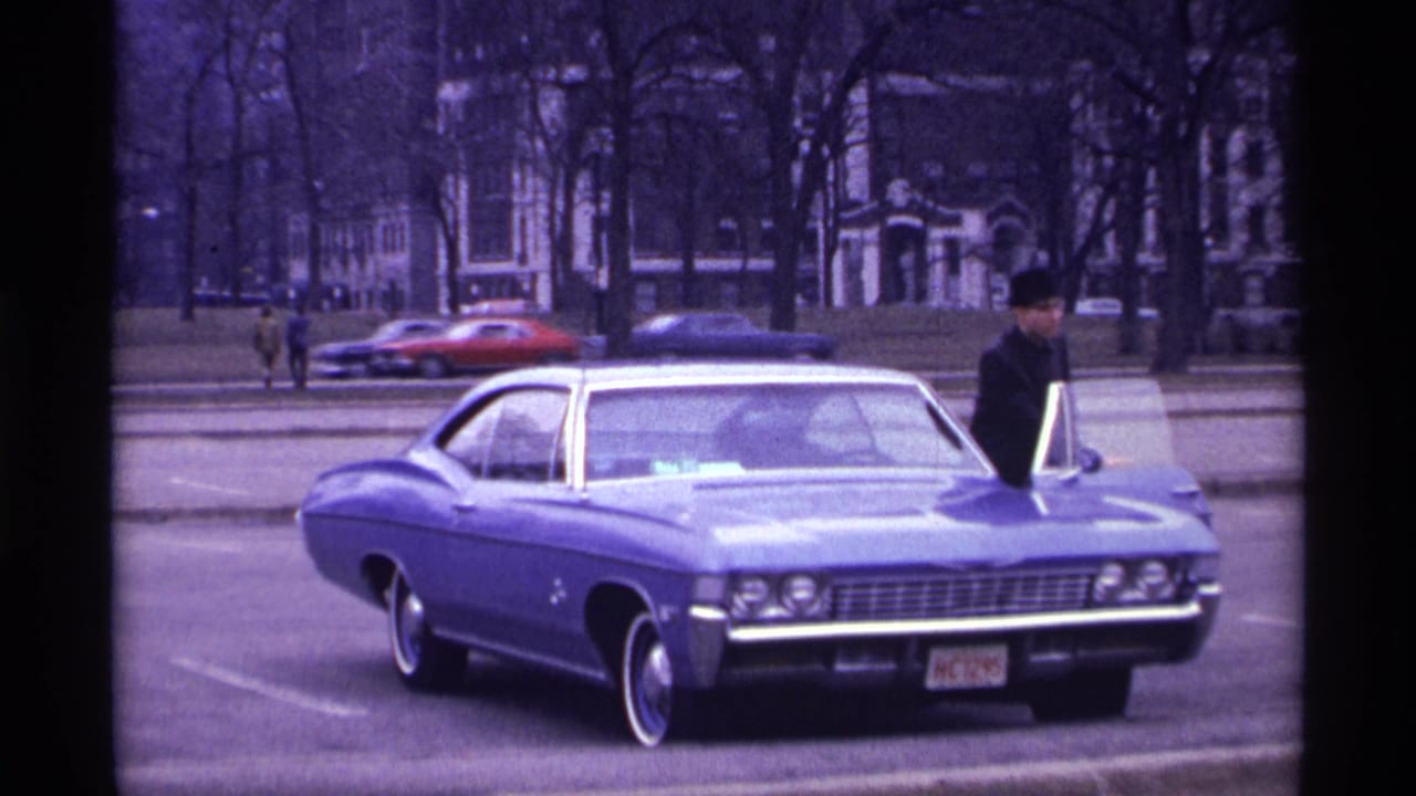 Blue classic car parked at curb with a man in a dark hat beside the open door.
