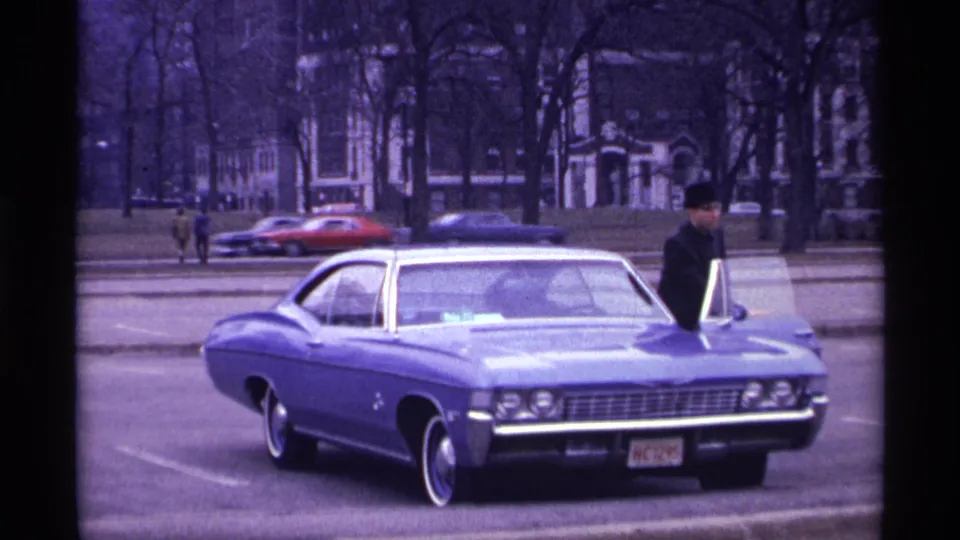 1968 Chicago home-movie frame showing a blue classic car at the curb with a suited man in a dark hat near the door.
