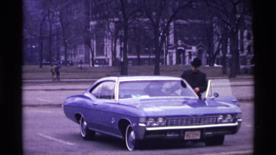 1968 Chicago frame of a blue classic car with chrome trim as a man in dark clothing enters or stands by the curbside door.
