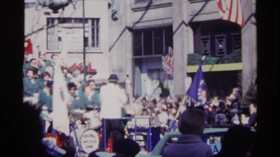 Curbside 8mm view of a Chicago parade float with a conductor, musicians in green uniforms, flags, and storefront signage.