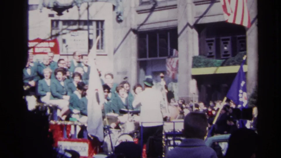 1968 Chicago parade frame showing a conductor in a white jacket facing a band on a float, with spectators and banners behind.