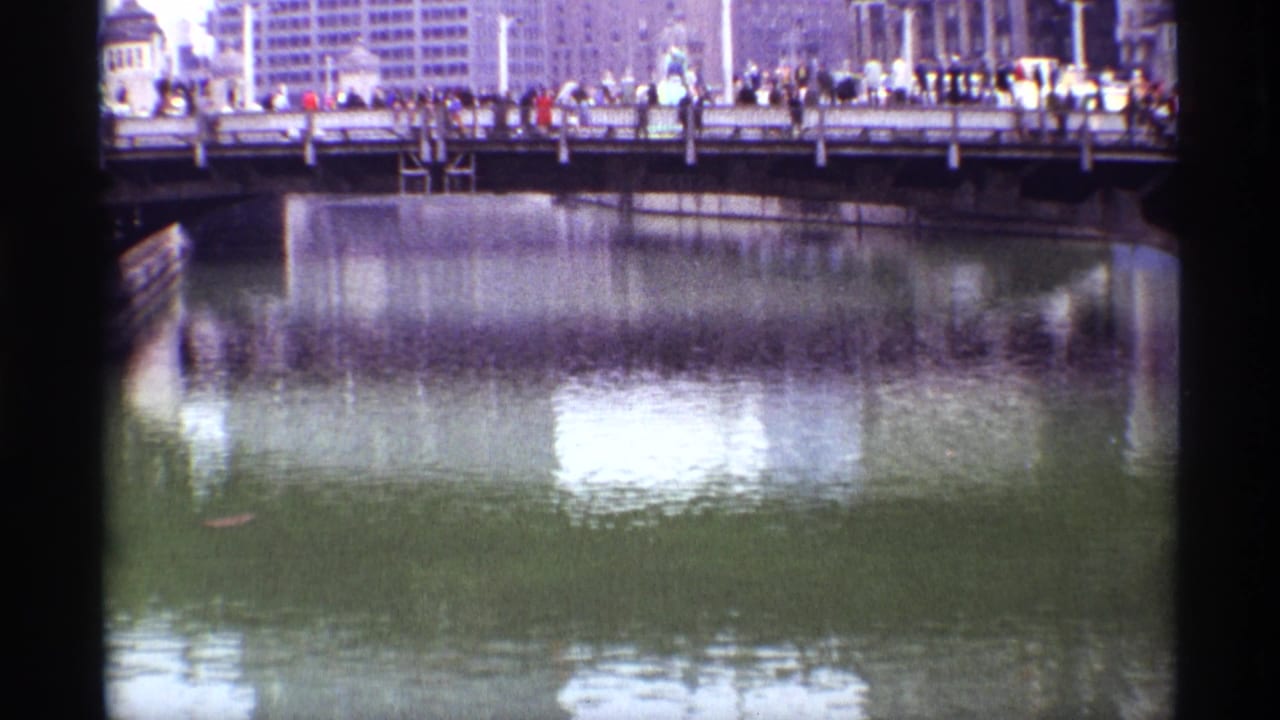 1968 Chicago riverfront frame with a crowded bridge railing, downtown buildings beyond it, and rippled reflections filling the foreground.
