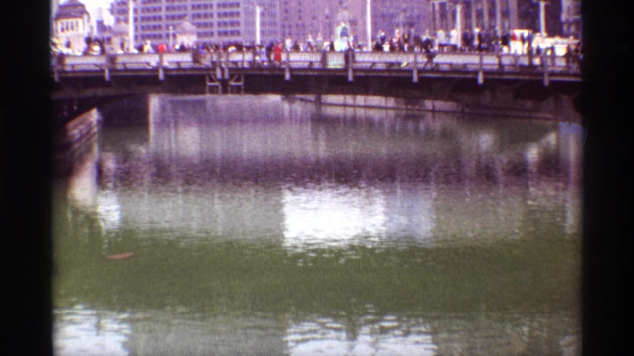 1968 Chicago downtown river scene with pedestrians on a bridge, vehicles near the railing, and broad building reflections across the water.
