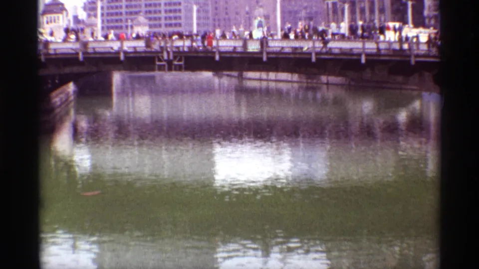 1968 Chicago downtown river scene with pedestrians on a bridge, vehicles near the railing, and broad building reflections across the water.