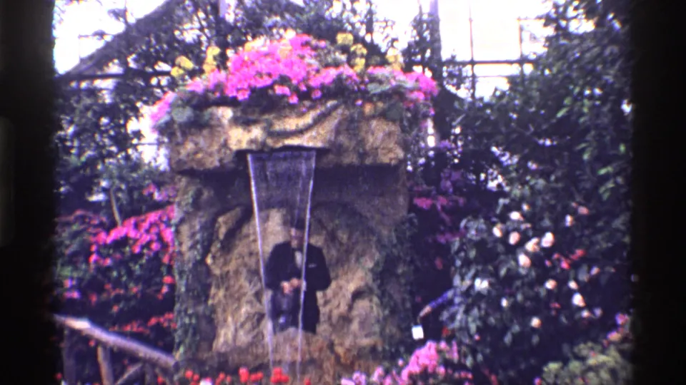 Soft 8mm frame from 1968 Chicago showing a suited visitor beneath a flowered fountain, partly shadowed by the water feature.