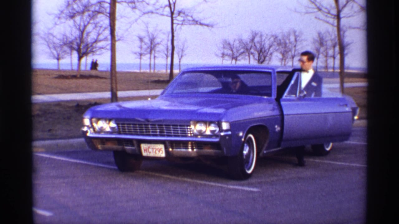 Blue classic sedan parked near bare trees with a man standing by the open driver-side door.