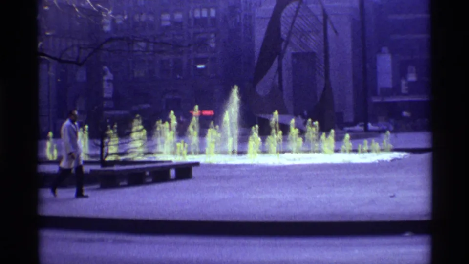 1968 Chicago plaza view with glowing fountain jets, bare trees, surrounding buildings, and a pedestrian crossing the foreground.