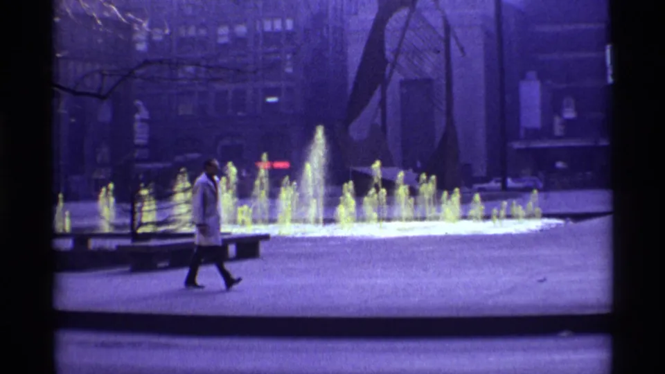 1968 Chicago frame of a man in a white coat crossing a public plaza, with glowing fountain jets and modern sculpture behind him.