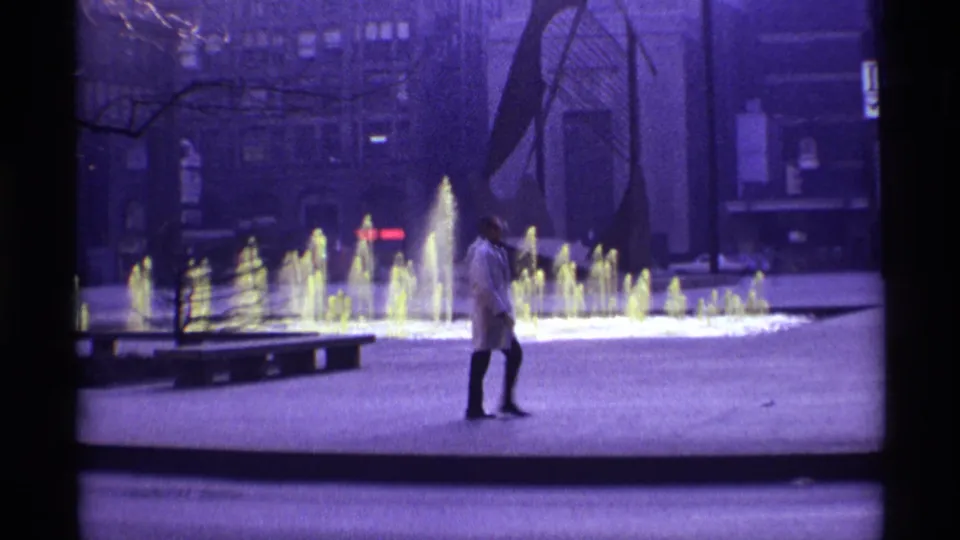 1968 Chicago home-movie frame showing a pedestrian in a white coat near bright fountain jets, bare trees, and a modern public sculpture.
