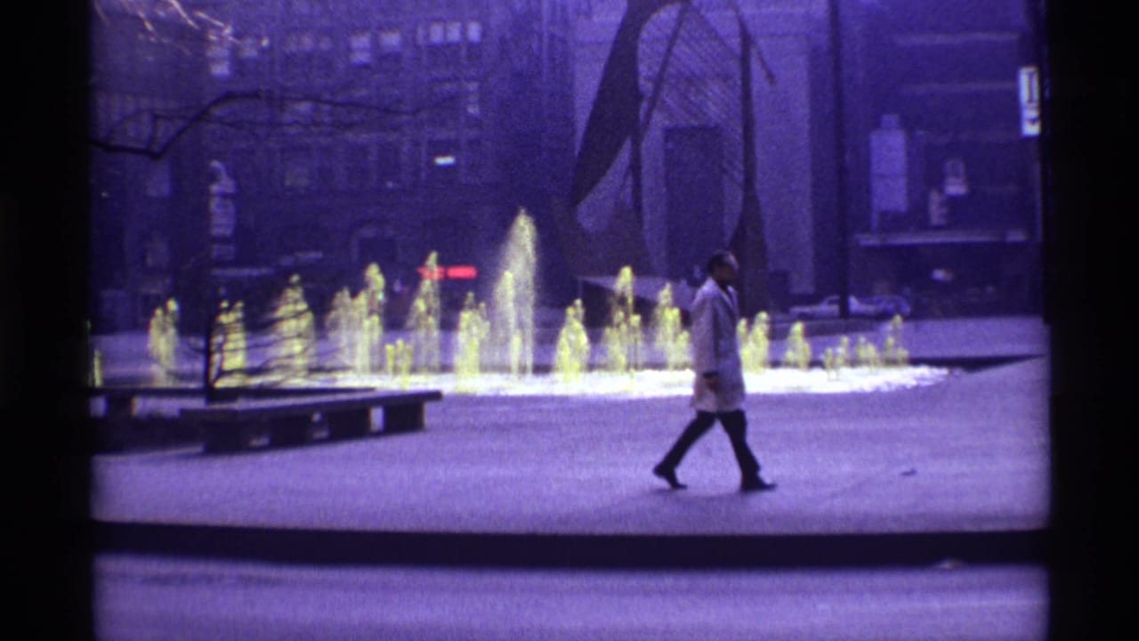 1968 Chicago home-movie frame showing a man crossing a paved plaza beside a glowing multi-jet fountain and bare trees.