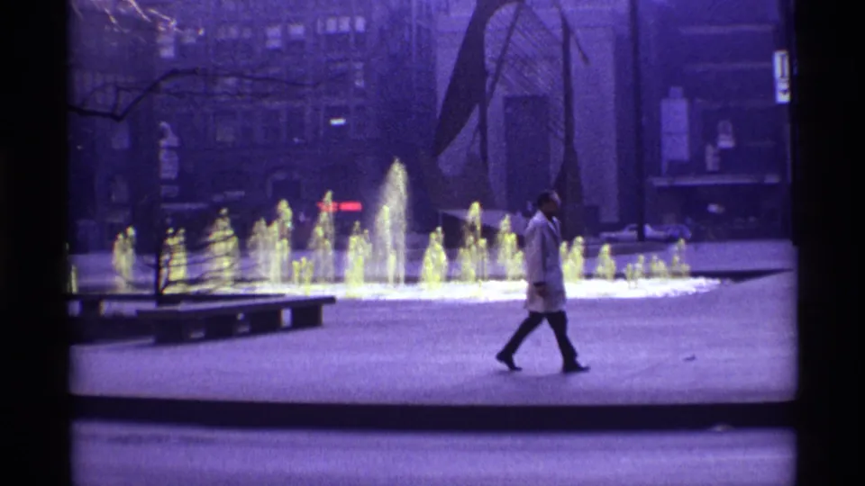 1968 Chicago home-movie frame showing a man crossing a paved plaza beside a glowing multi-jet fountain and bare trees.