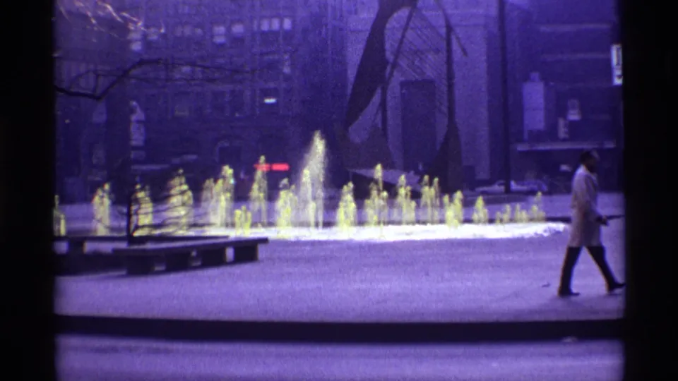 Chicago plaza scene with yellow-lit fountain jets, bare trees, and a man in a light coat moving out of frame.