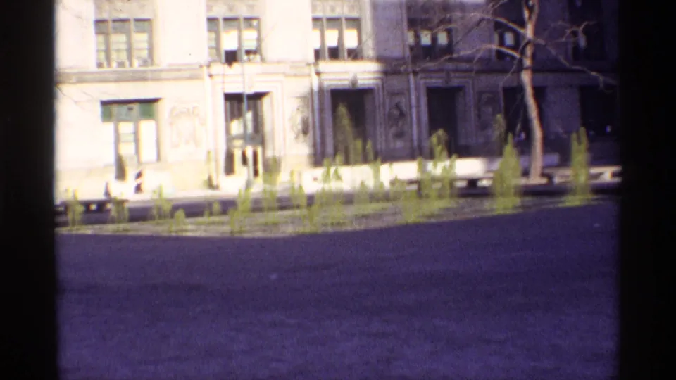 Brighter 8mm frame of the Chicago square showing fountain jets, bare trees, benches, and building facades.