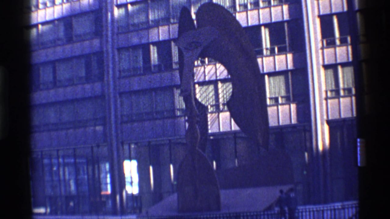 Abstract sculpture in front of a gridded urban building with small pedestrians nearby.