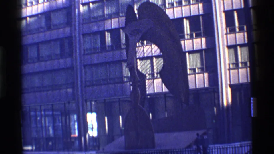 1968 Chicago home-movie frame showing a large abstract public sculpture against a modern gridded building facade with distant pedestrians.