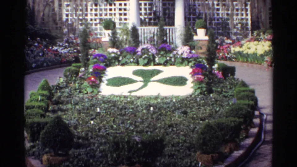 Chicago garden scene with shaped hedges, colorful flower beds, paved paths, and downtown buildings in the background.