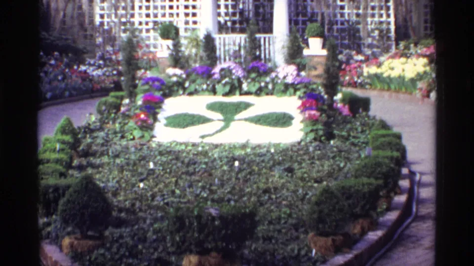 Chicago garden cutaway with dense hedges, colorful flower beds, paved paths, and buildings visible beyond the plantings.