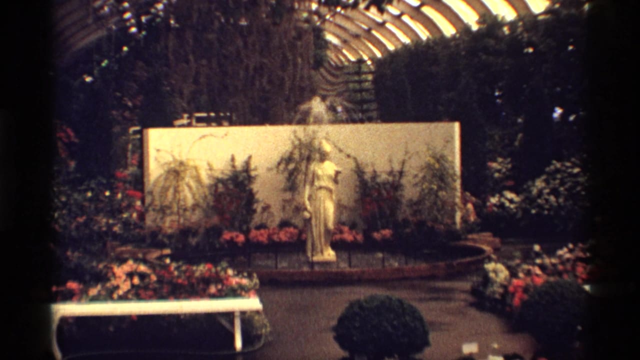 Greenhouse display with a female statue, fountain spray, flowers, and a curved glass roof.