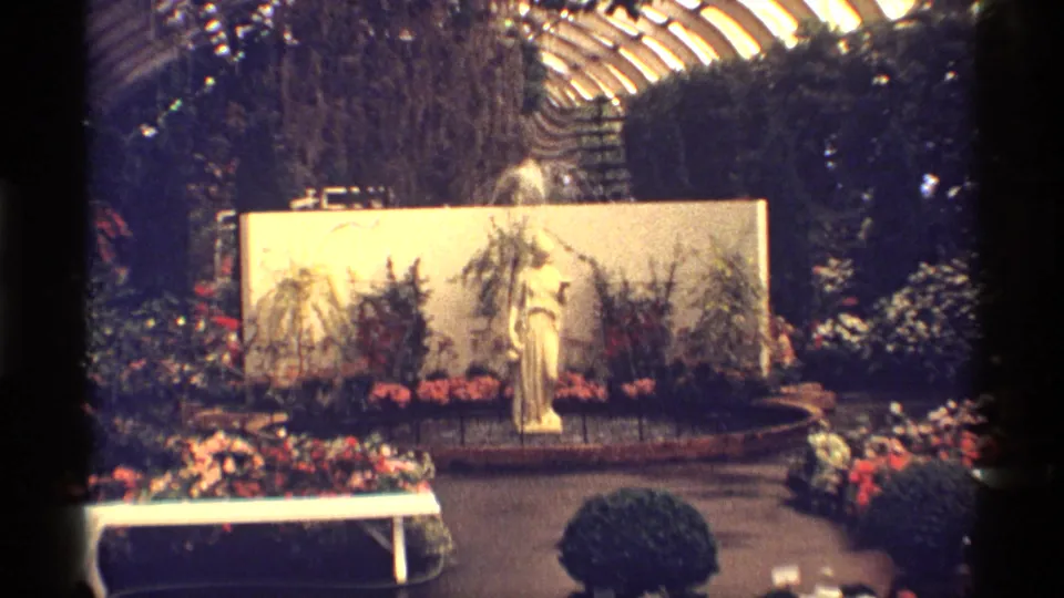 1968 Chicago 8mm still with a greenhouse statue, fountain spray, arranged flowers, a low white bench or rail, and plants beneath the glass ceiling.