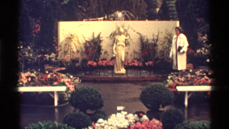 Wide 1968 Chicago garden view with a statue, surrounding flower beds, benches, shrubs, and a woman at the right.