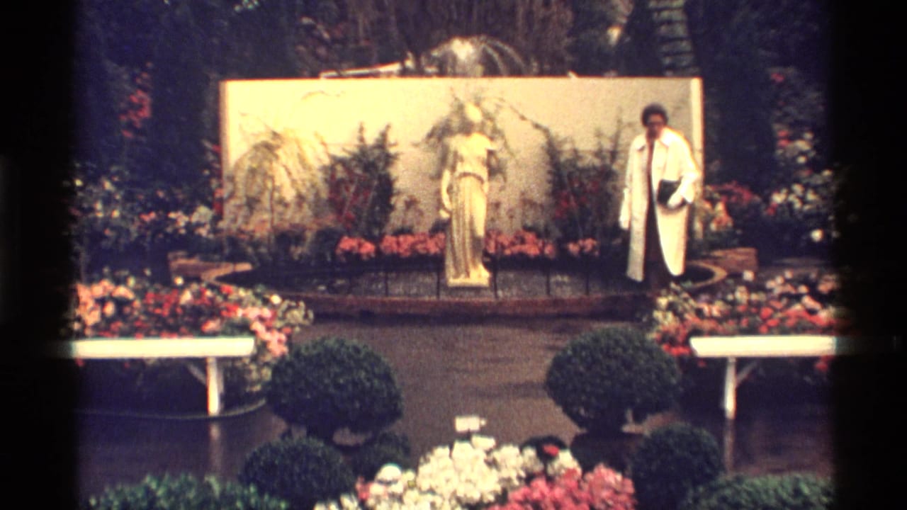 In 1968 Chicago home-movie footage, a woman in a white coat walks past flowers, shrubs, benches, and a central statue.