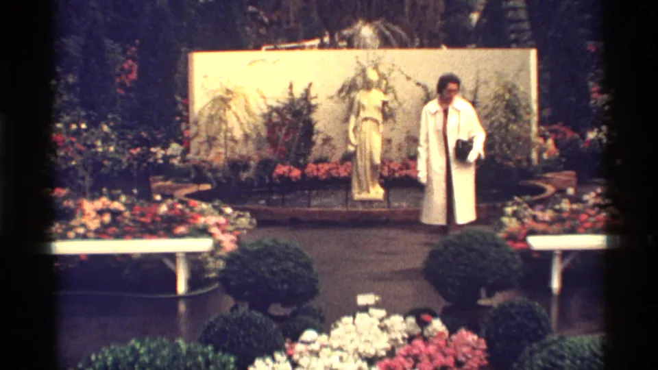 A woman in a white coat faces the camera in a colorful 1968 Chicago garden scene with a statue, benches, shrubs, and flower beds.