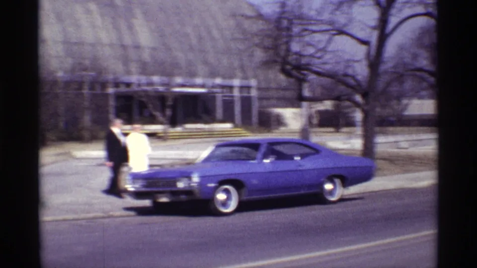 A man in a dark suit and woman in a light dress stand by the front of a purple classic car in Chicago home-movie footage.