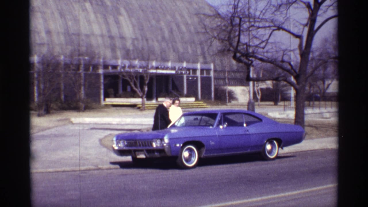 Suited man and woman stand beside a purple two-door car on a city street.