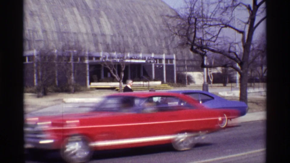 A red car blurs across the foreground, partly obscuring the suited man and purple coupe in the 1968 Chicago street scene.
