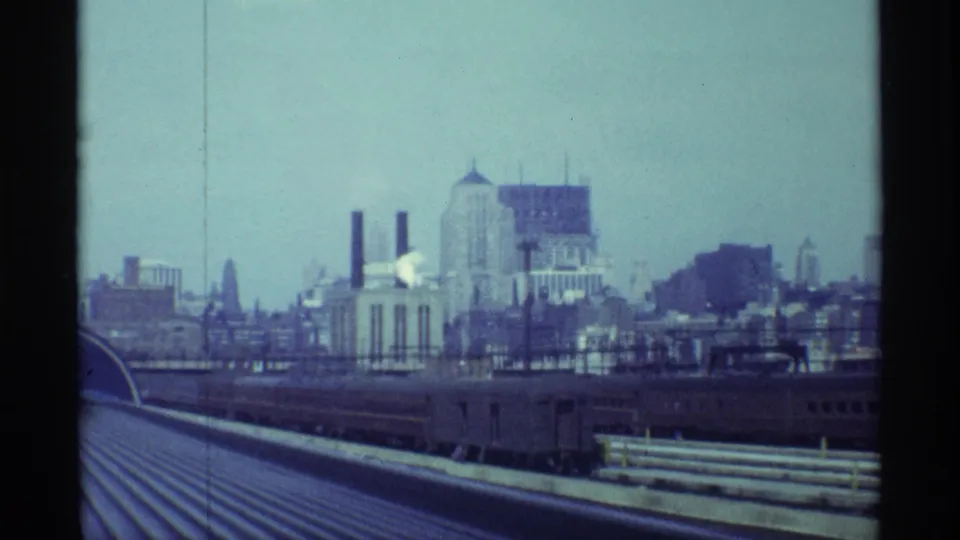 Chicago rail approach from above a train, with the car roof leading toward industrial structures, smokestacks, and distant skyline forms.
