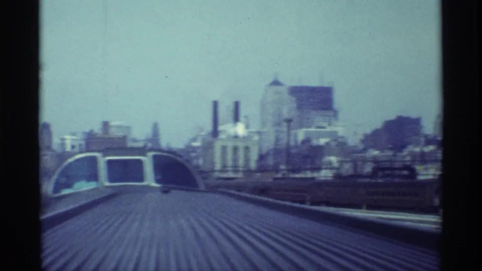 Chicago train-top view approaching an arched rail or station structure, with industrial buildings, smokestacks, and skyline visible beyond.