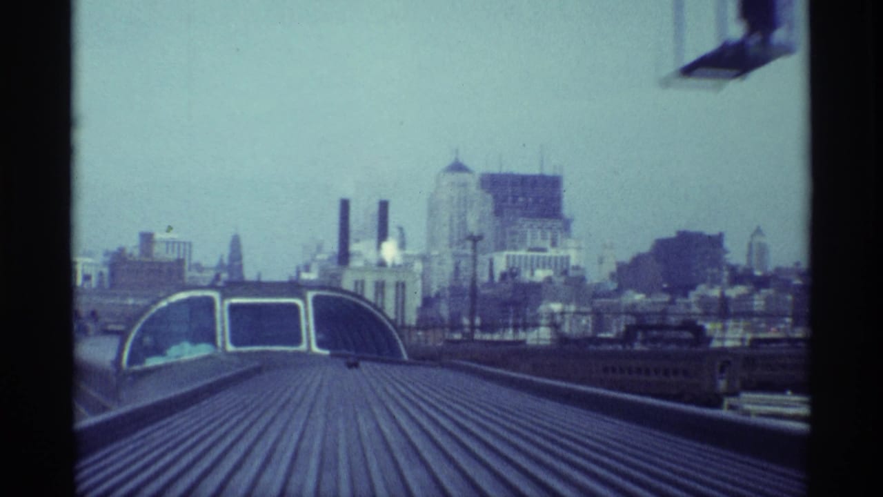 Train roof view toward a hazy urban skyline with smokestacks and industrial buildings.