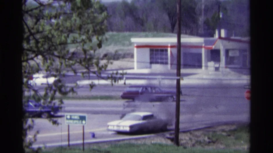 1968 Chicago roadside view with passing cars, trees, utility poles, and a white-and-red station building in the background.