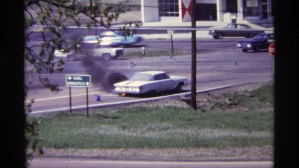 White car burning along a Chicago roadside in 1968, with dark smoke, passing cars, signage, grass, and a station building visible.