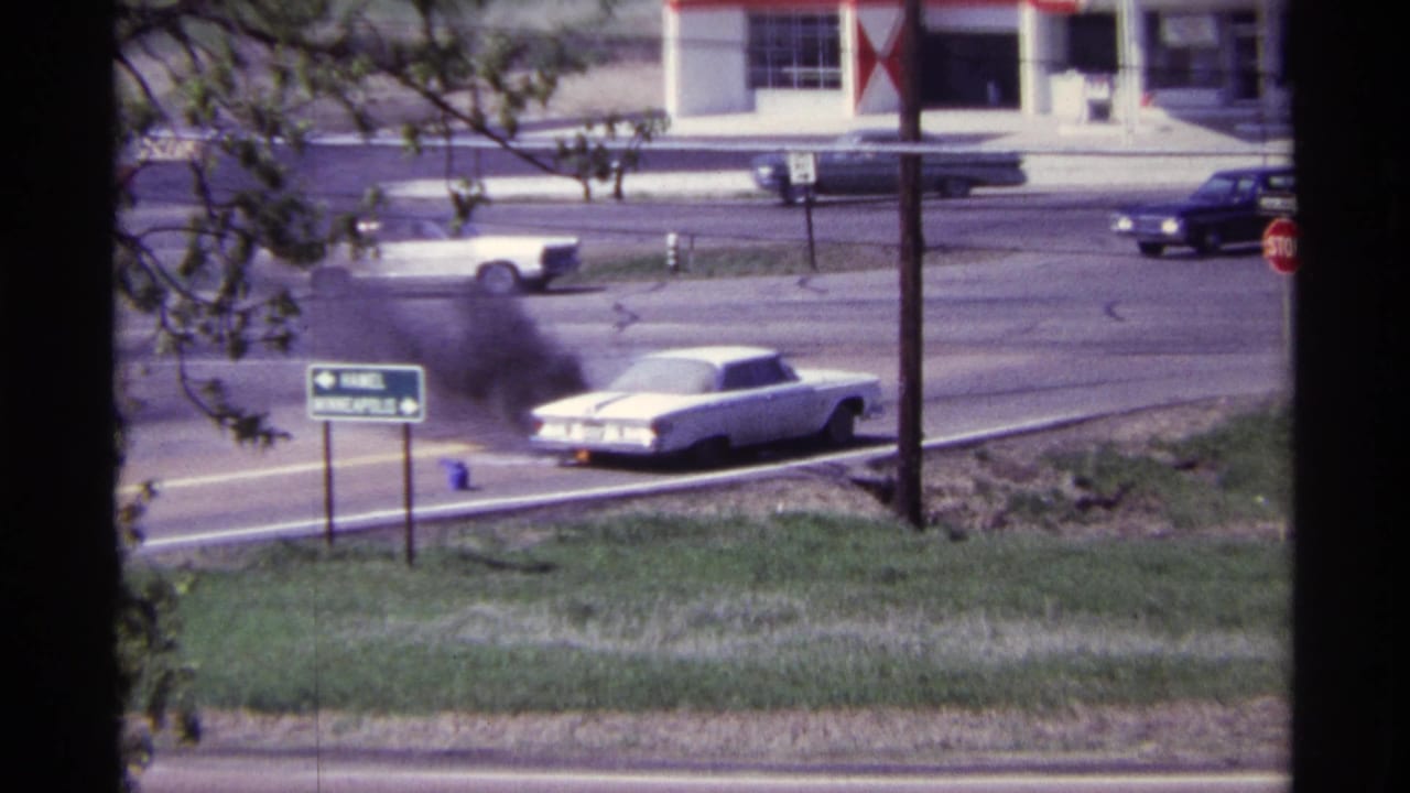 1968 Chicago-area 8mm frame of a white classic car smoking heavily beside a suburban road near a service-station facade.