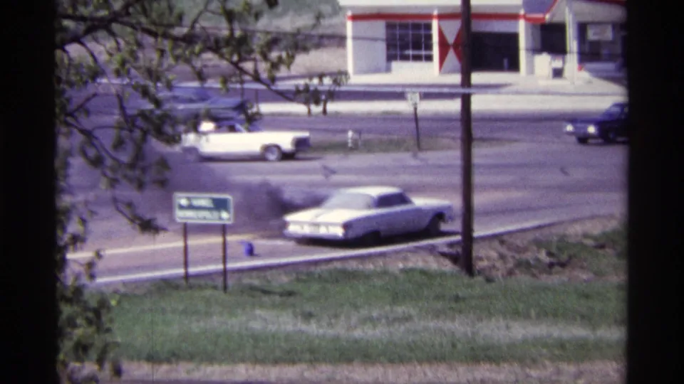 The white car continues to emit black smoke on a Chicago-area suburban road, with period vehicles passing behind it.
