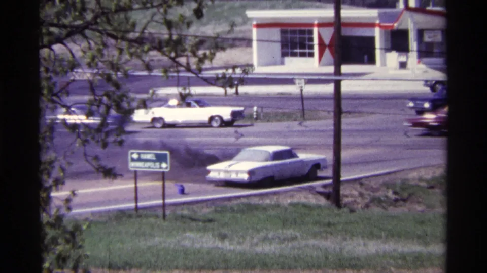 A 1968 roadside view shows the smoking white car in the foreground with multiple period vehicles moving past a gas station.