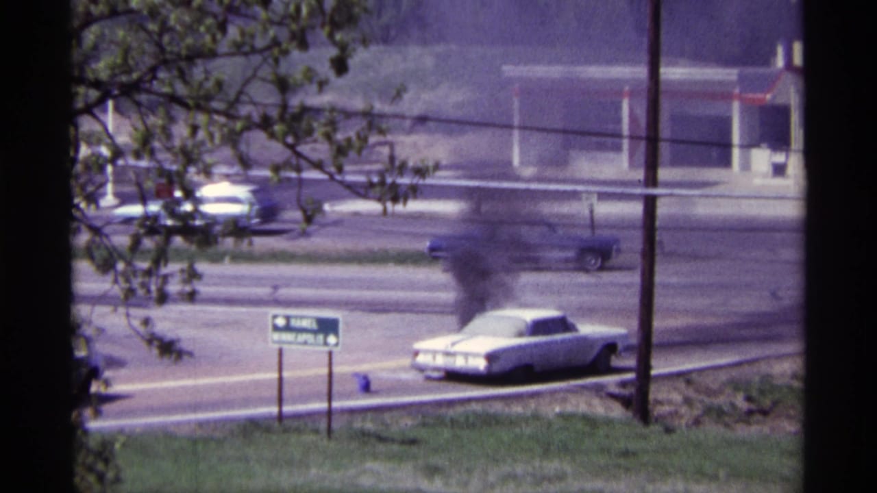 White car smoking beside a road with traffic, trees, signs, and a commercial building nearby.