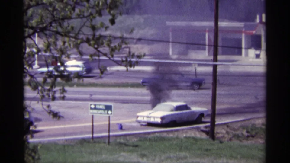 1968 Chicago roadside view of a white car emitting dark smoke as traffic passes near signs and a low commercial building.