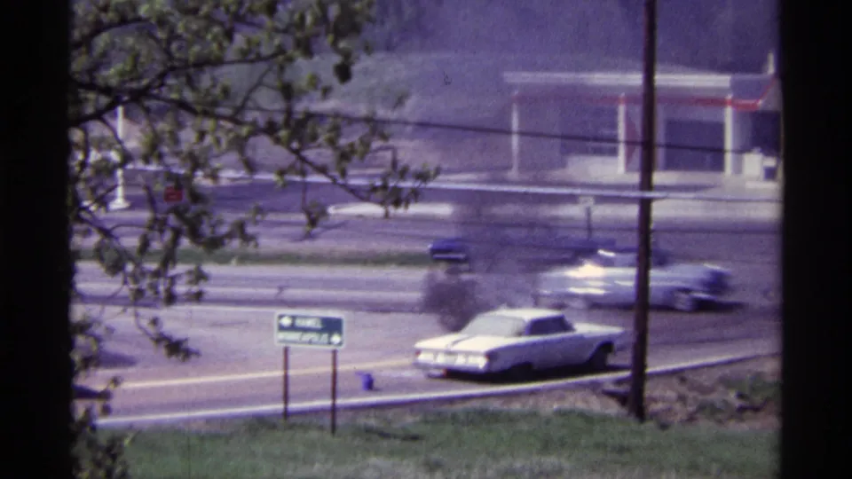 1968 Chicago traffic passes a white roadside car releasing smoke near a sign, trees, utility poles, and a low building.