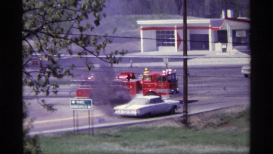 A red fire truck reaches the smoking white car on a Chicago roadside in the supplied 1968 home-movie footage.
