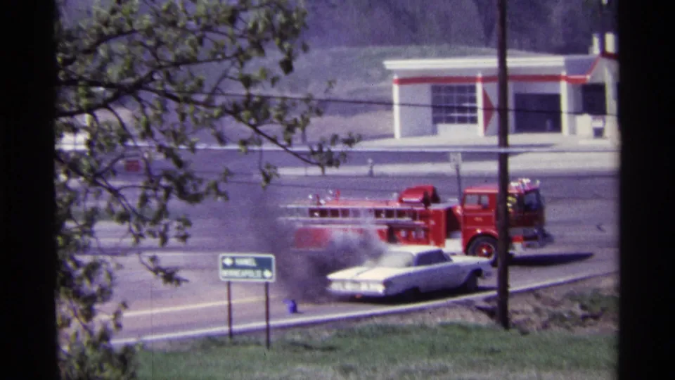 Red fire truck positioned next to the smoking white car on a Chicago roadway, with signs, branches, and a commercial building visible.