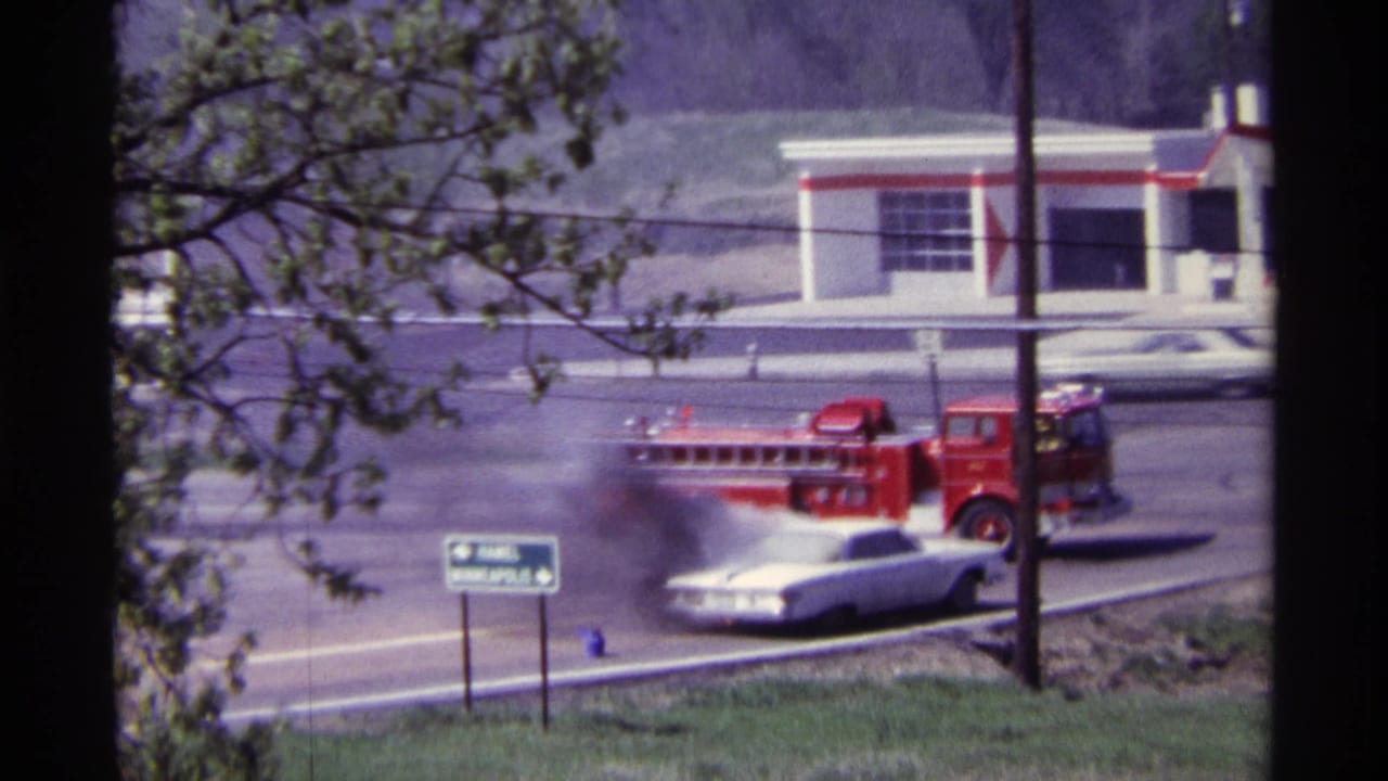 White sedan smoking at roadside as a red fire truck stops nearby.